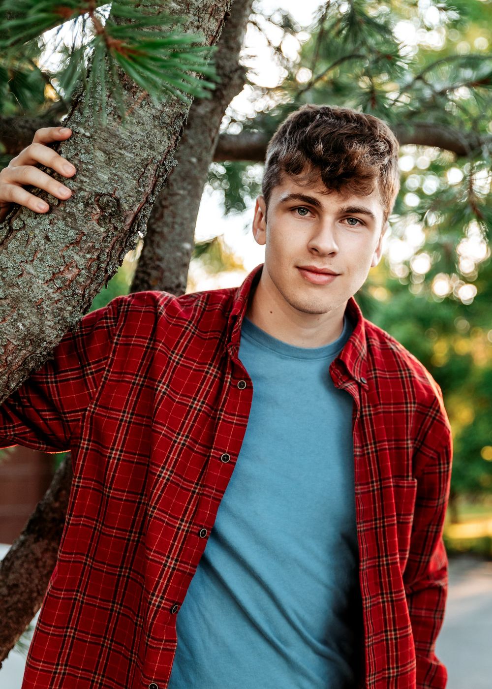 A person wearing a red plaid shirt and blue t-shirt poses casually against a tree trunk in an outdoor setting.
