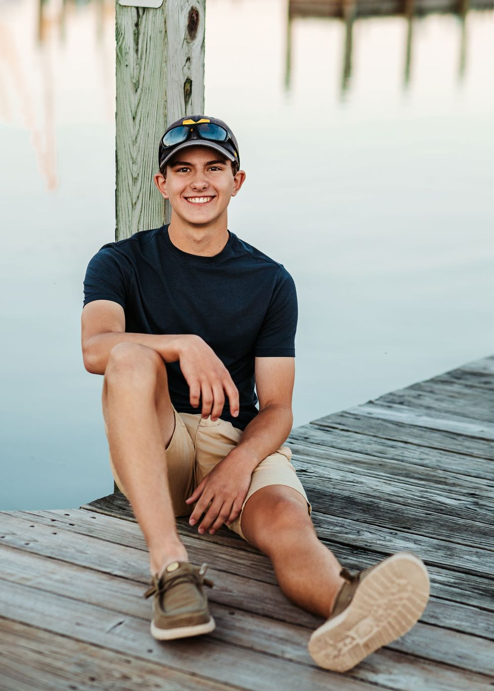 A person in a black t-shirt and khaki shorts sits casually on a wooden dock on a bright sunny day.
