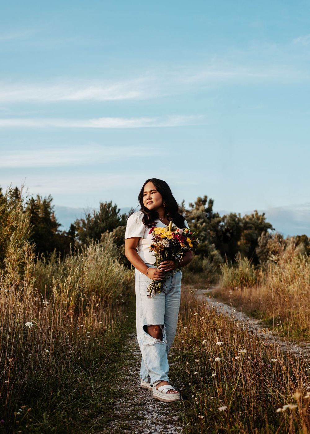 A path winds through golden sunlit wildflowers and tall grass against a bright blue sky during sunset.