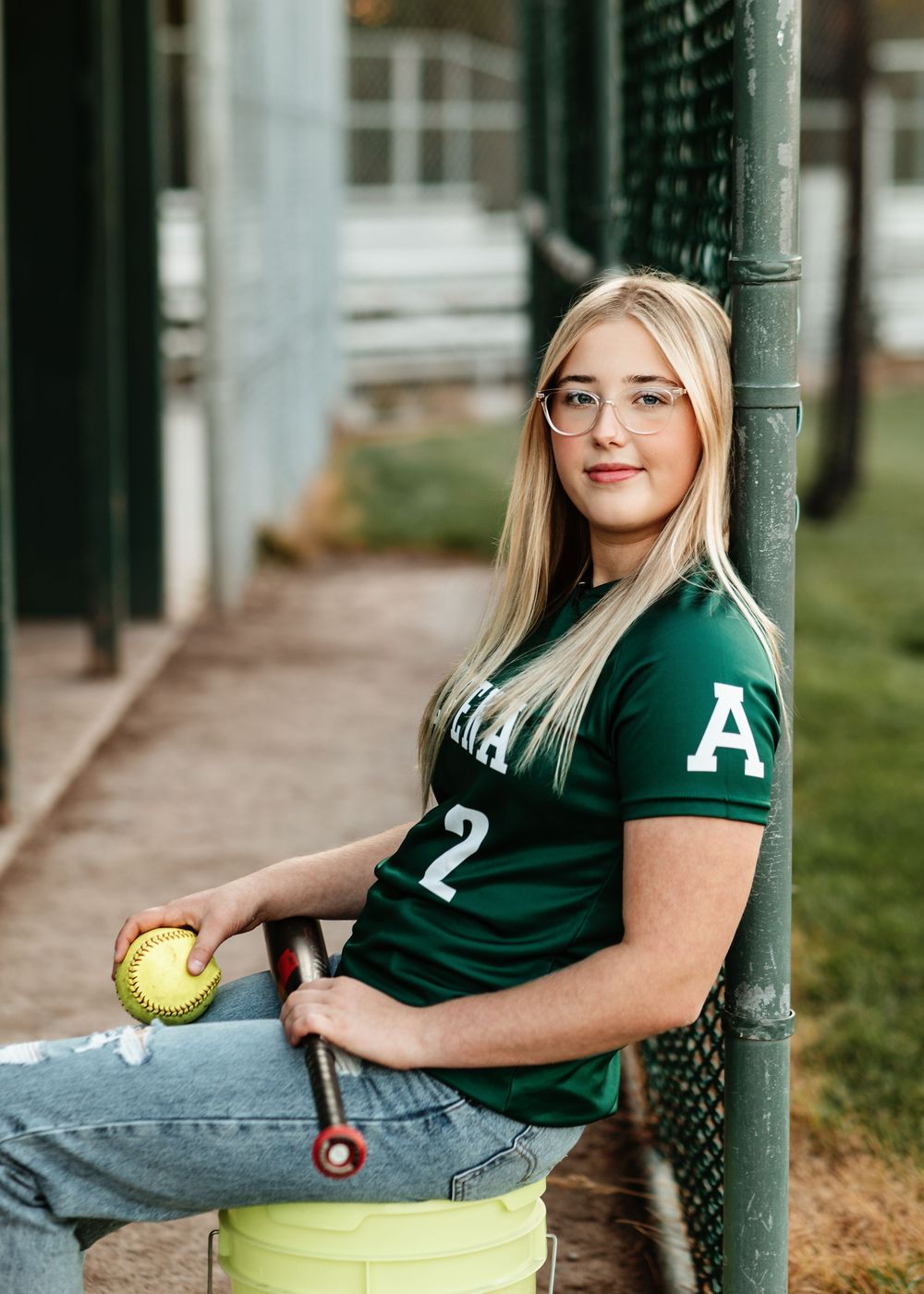 A softball player in a green jersey and jeans sits on a metal pole while holding a yellow ball outdoors.