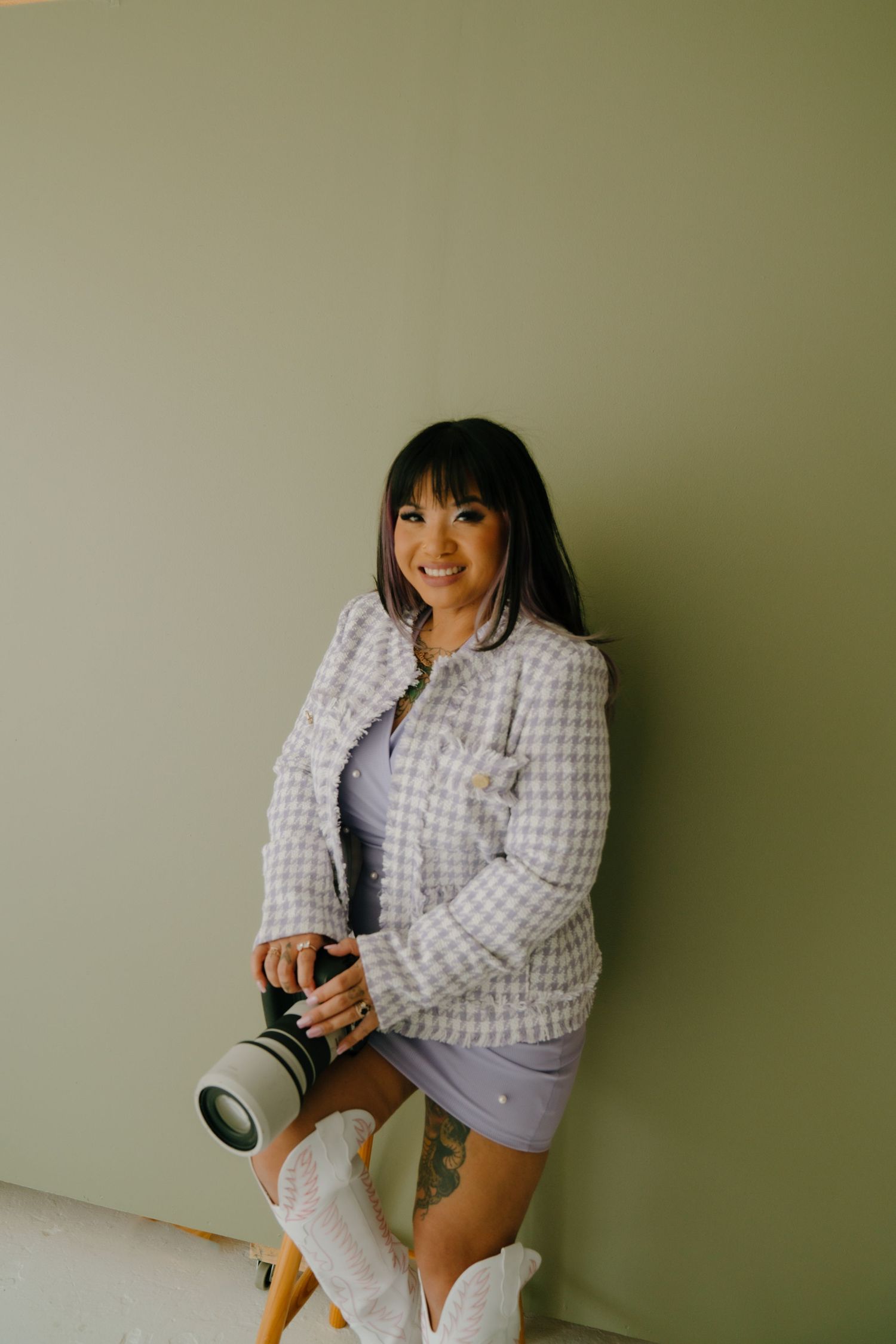 Professional photographer in casual attire posing with camera equipment against neutral wall.