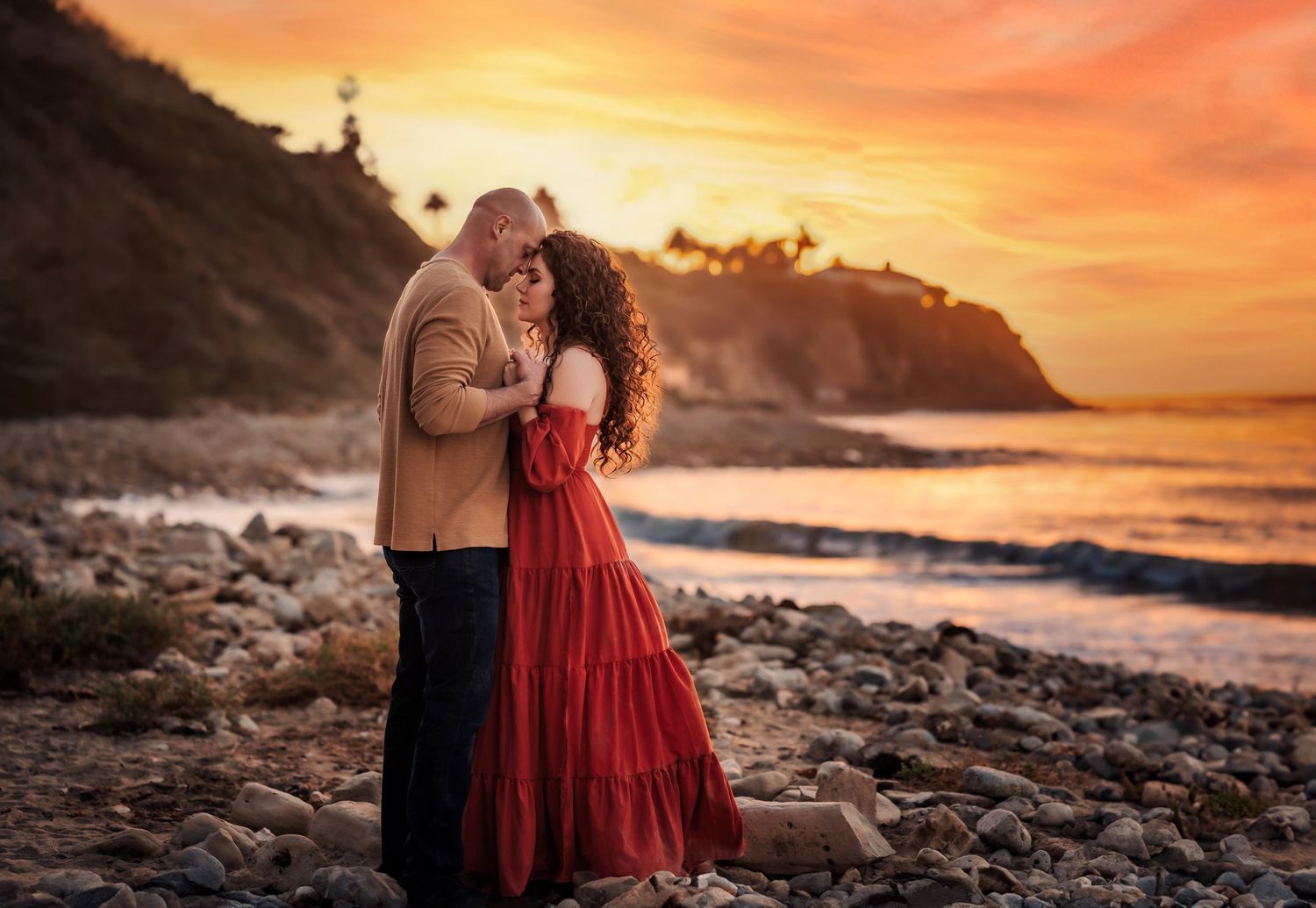 Couple sharing romantic moment on rocky beach during dramatic orange sunset.