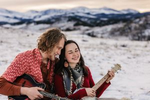 Couple playing ukulele together in snowy mountain landscape at dusk, smiling and sharing a joyful musical moment.