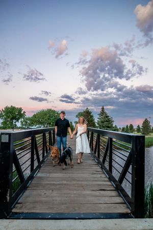 Couple walking two dogs on a wooden bridge at sunset with colorful sky and green trees in background.