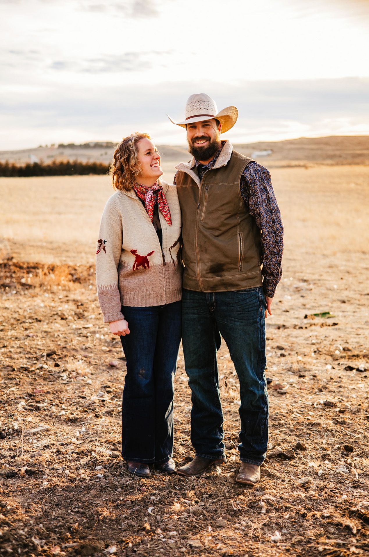 Couple stands together in western attire on a golden field at sunset with mountains in the background.
