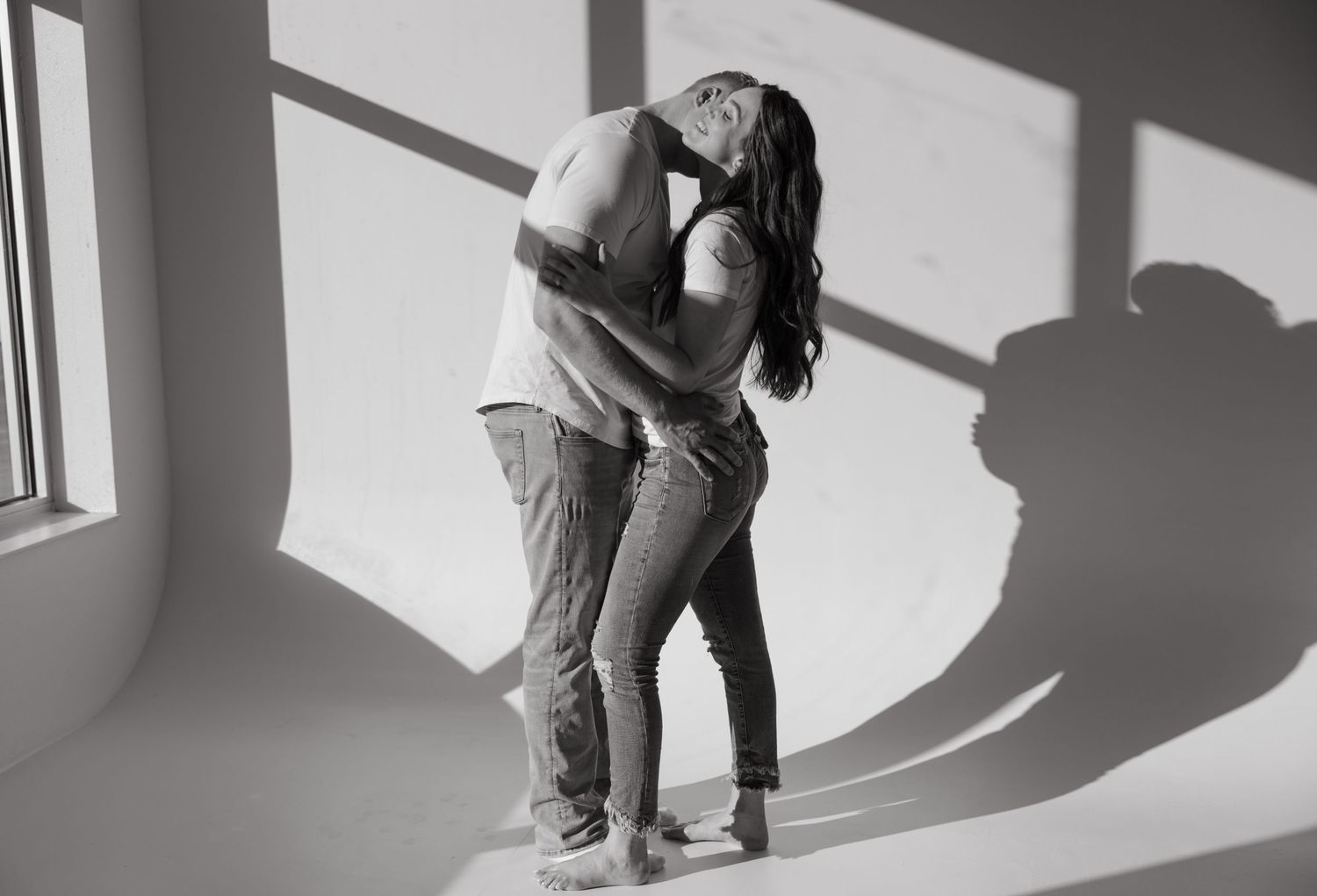 A couple embraces in casual clothing casting dramatic shadows in a minimal white room.