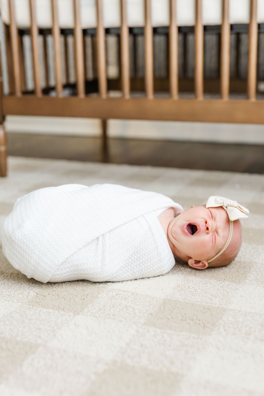 Swaddled newborn baby girl with bow headband yawns while lying on floor near wooden crib.
