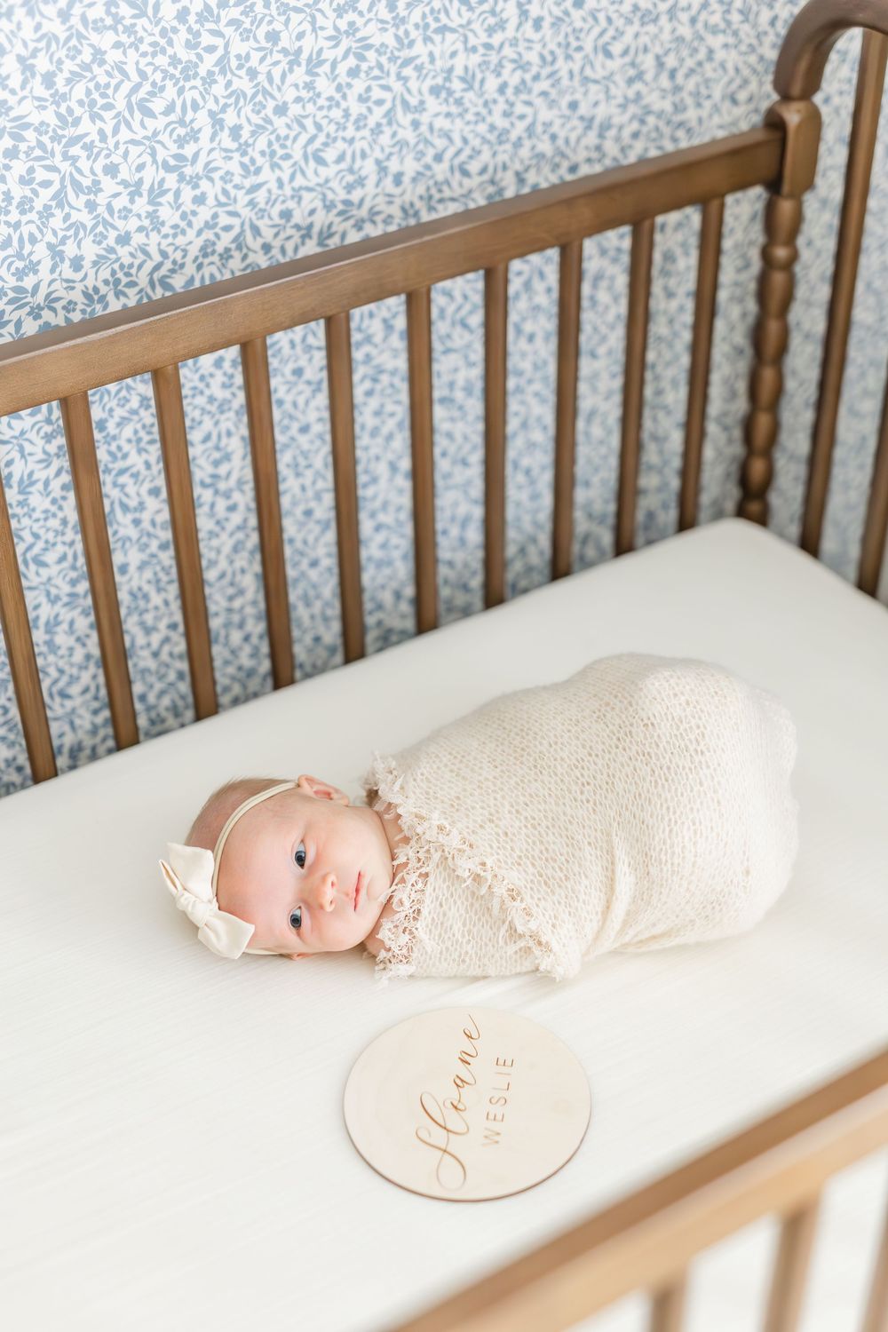 Newborn baby girl swaddled in cream wrap lies in wooden crib with name disc milestone marker.