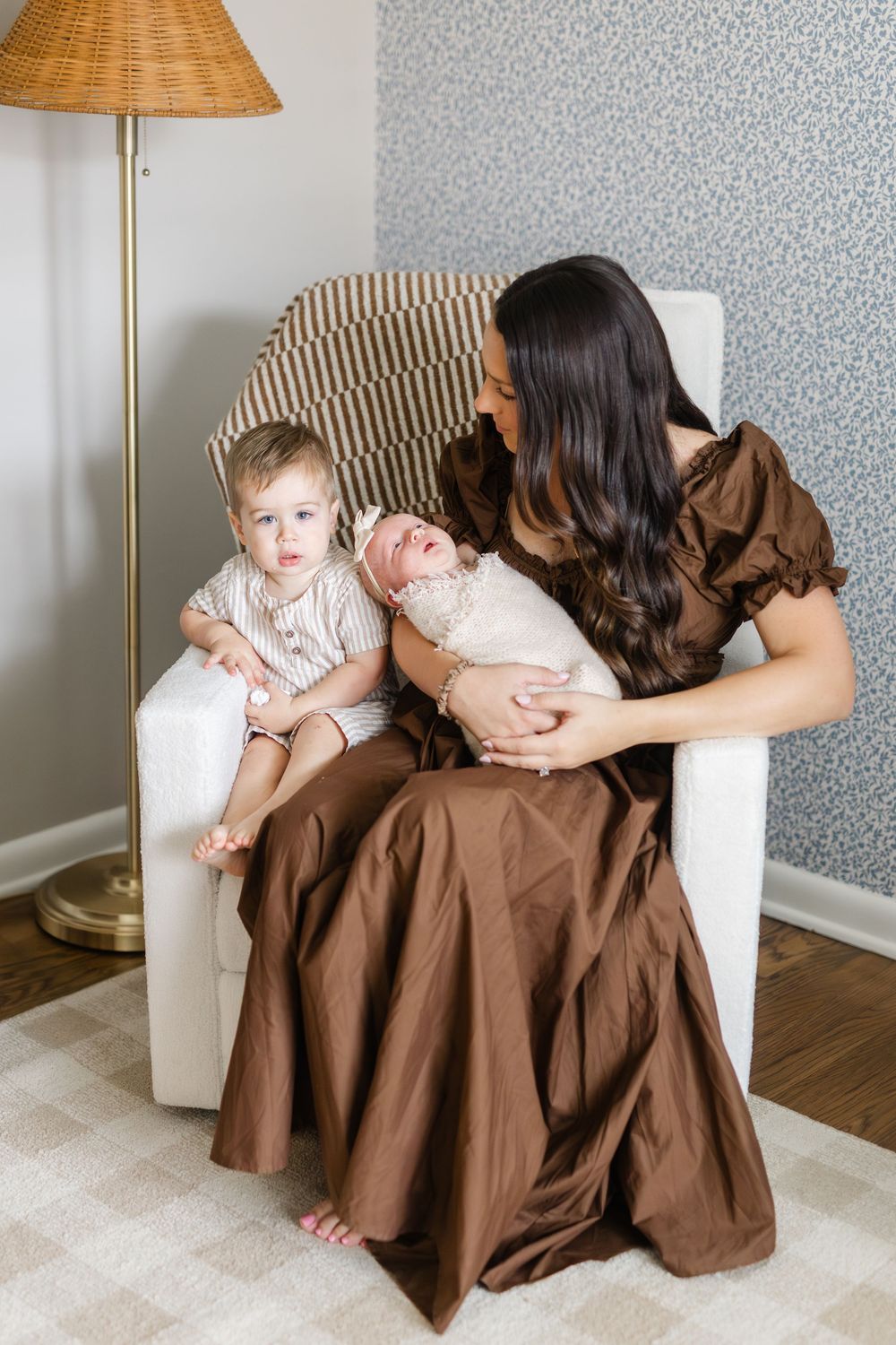Mother in brown dress sits in nursery chair holding newborn while toddler son sits beside her.