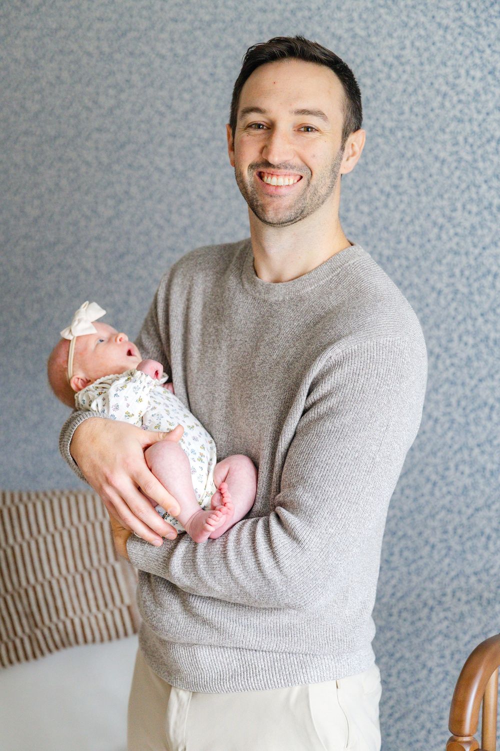 Smiling father in gray sweater proudly holds his newborn baby girl in floral outfit.