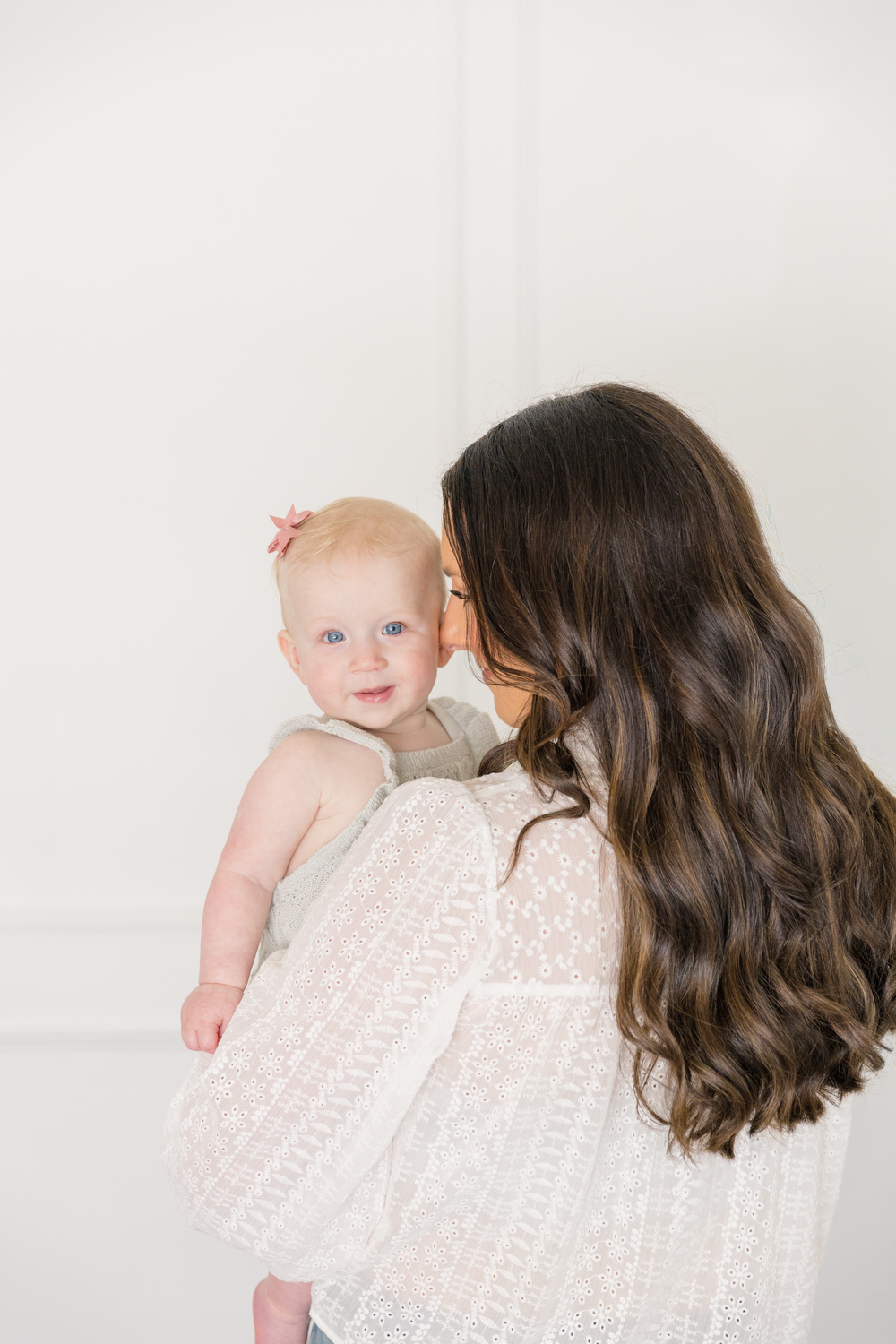 A mother with long dark hair kisses her smiling baby girl in a bright white room.