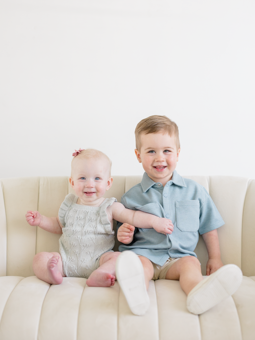 Two smiling siblings, a baby girl in gray romper and toddler boy in blue outfit, sitting together on a cream sofa.