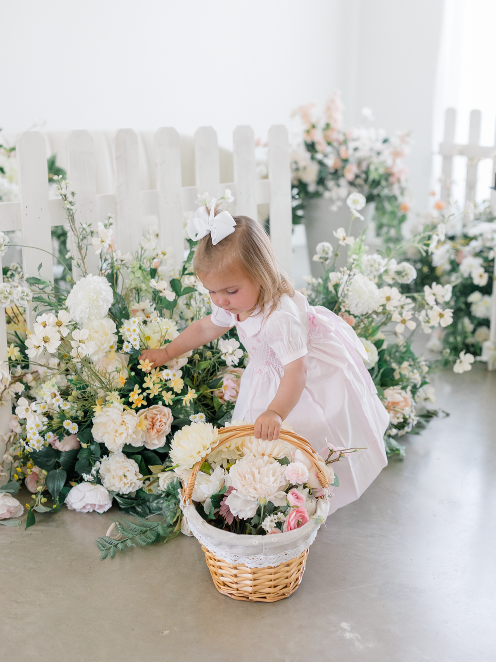 Young girl in pink dress picking flowers from a basket near a white picket fence surrounded by blooms.