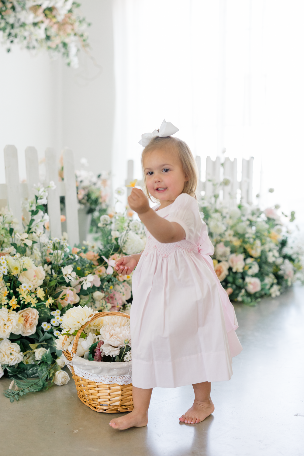 Toddler girl in pink dress with white bow holds Easter basket among spring flowers.