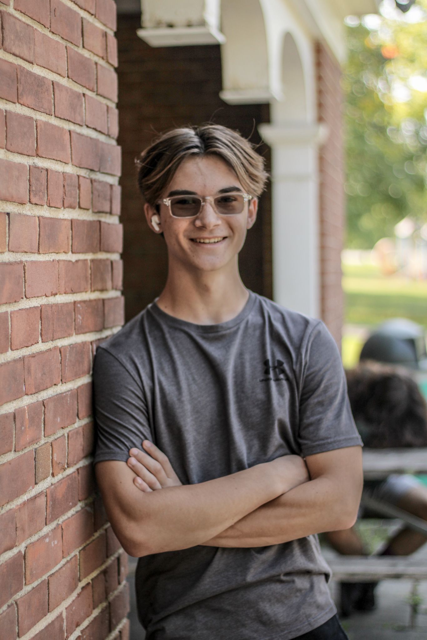 Person in grey shirt leaning against brick wall wearing sunglasses.