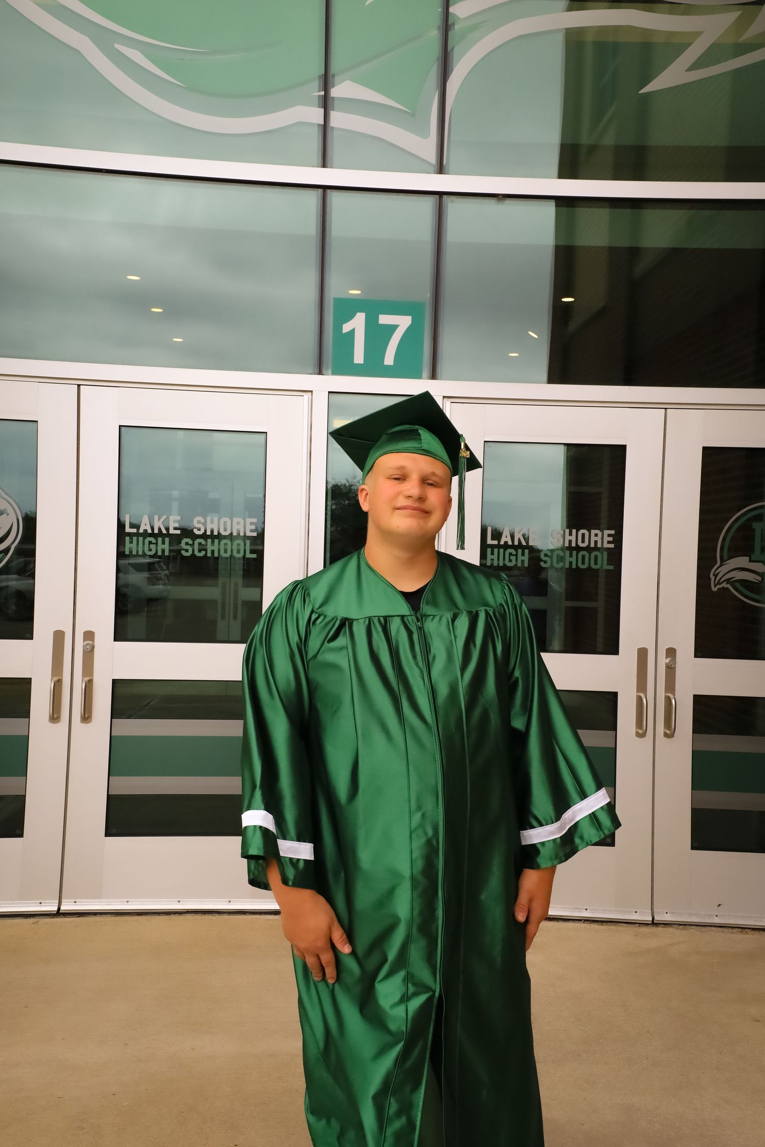 Graduate in green regalia standing in front of school entrance labeled 17.