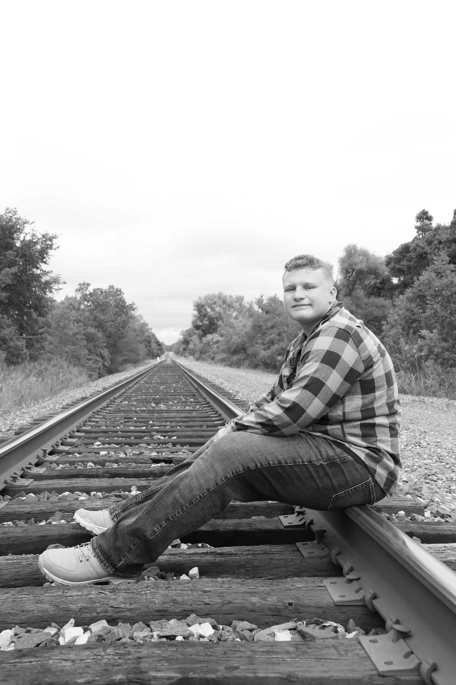 Black and white portrait of person sitting on railroad tracks wearing plaid shirt and jeans.