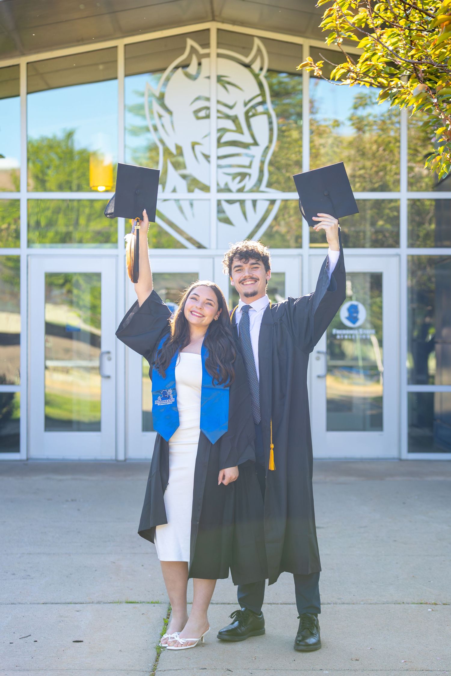 Two graduates in black gowns raise their caps in front of a school building with a blue devil mascot logo.