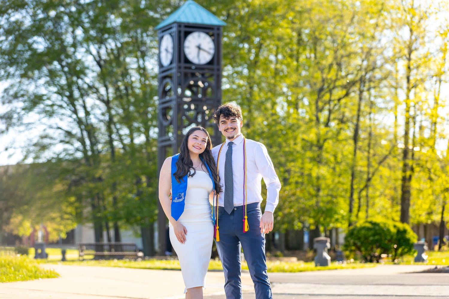 Graduating couple poses in front of a clock tower on a sunny spring day on a college campus.