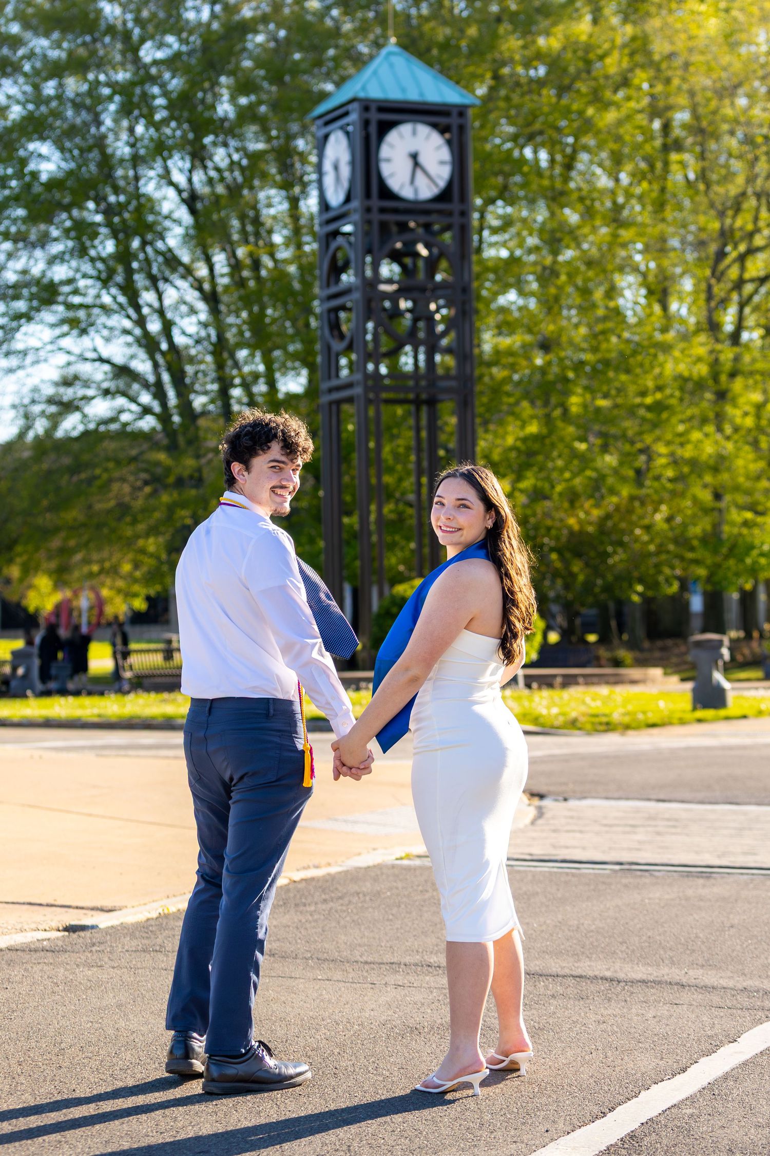 A couple holds hands and smiles near a clock tower surrounded by green trees on a sunny day.