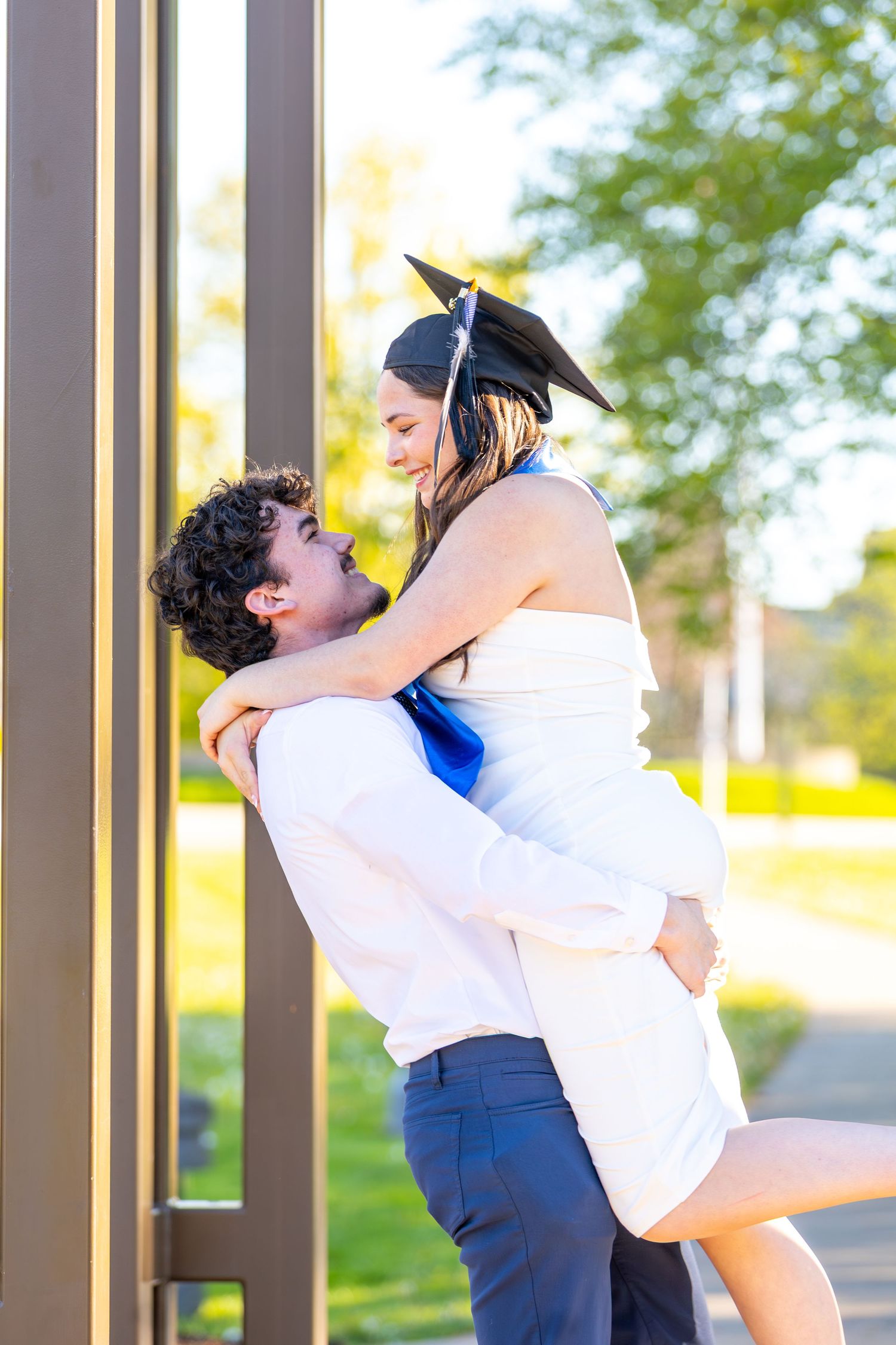 A man lifts a smiling woman in a graduation cap and white dress, celebrating her achievement outdoors.