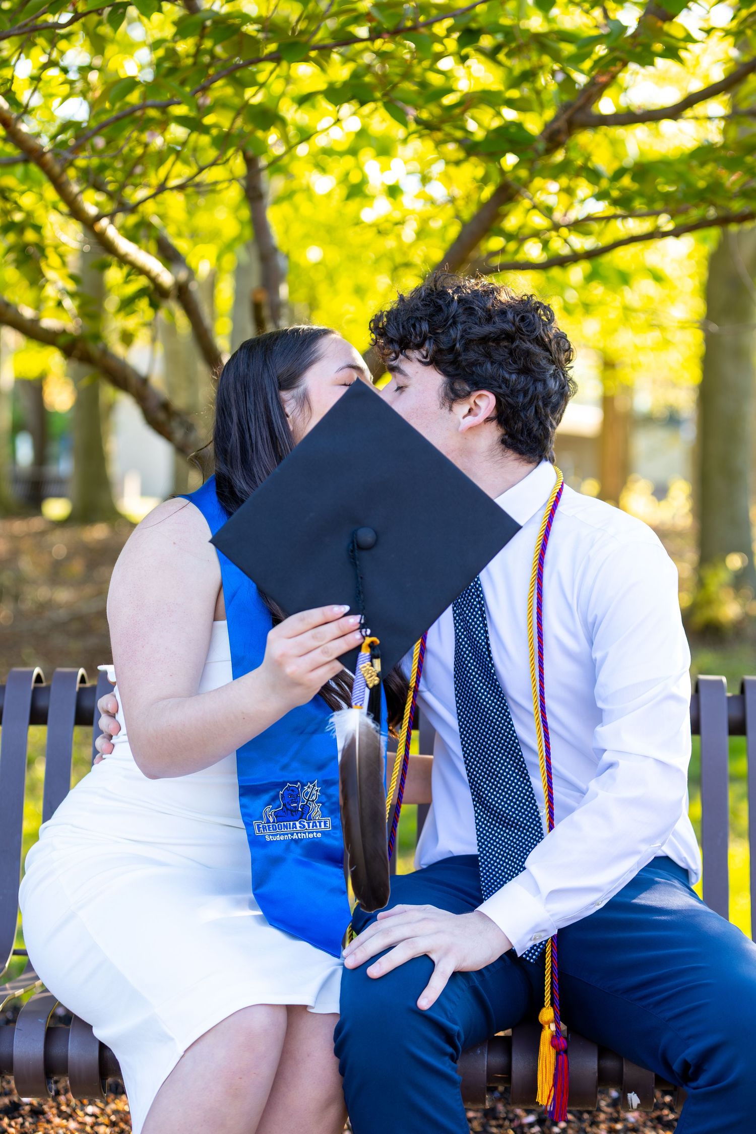 Couple sharing a kiss behind a graduation cap on a bench surrounded by autumn trees.