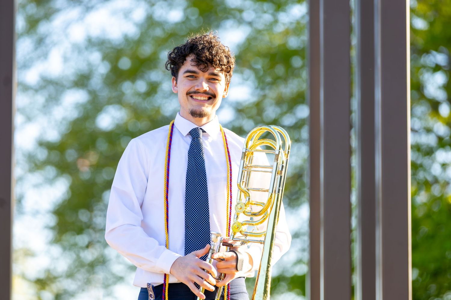 Young man in white shirt and tie holds a trombone outdoors, wearing orange graduation stole.