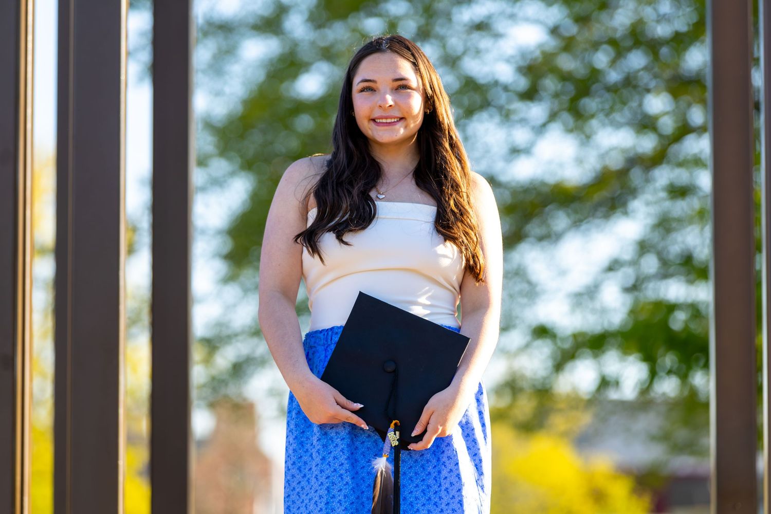 Smiling young woman in white top and blue polka dot skirt holding graduation cap outdoors on a sunny day.