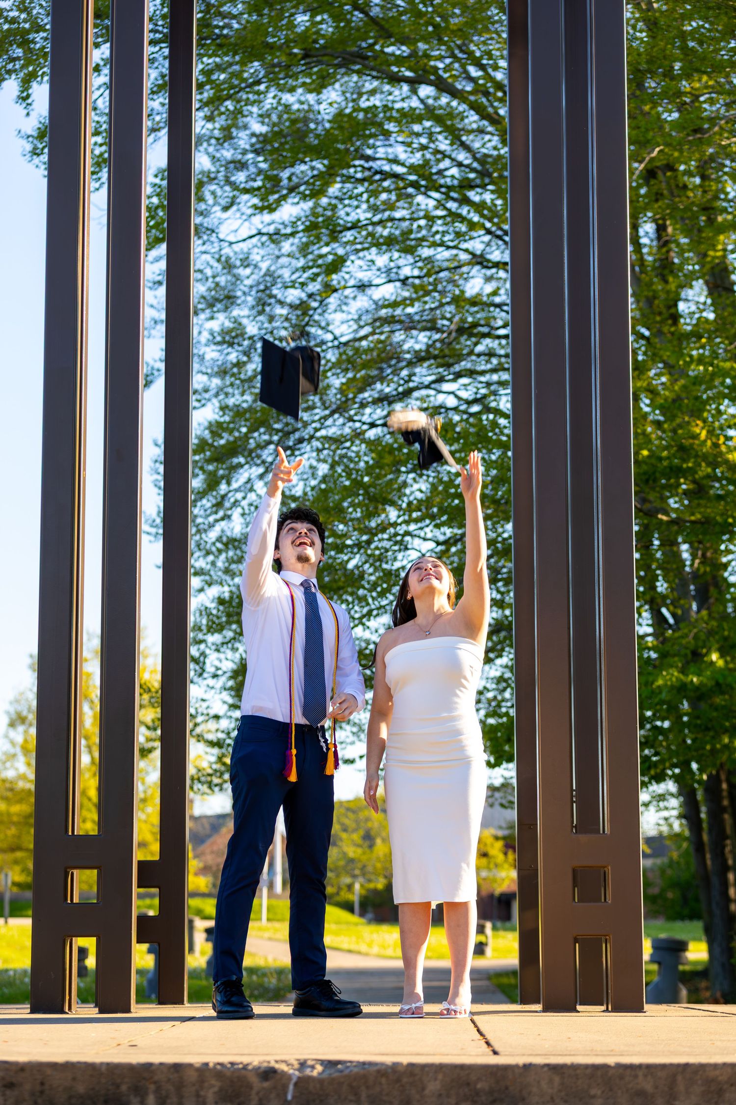 Two graduates joyfully tossing caps in the air outdoors near metal sculptures.