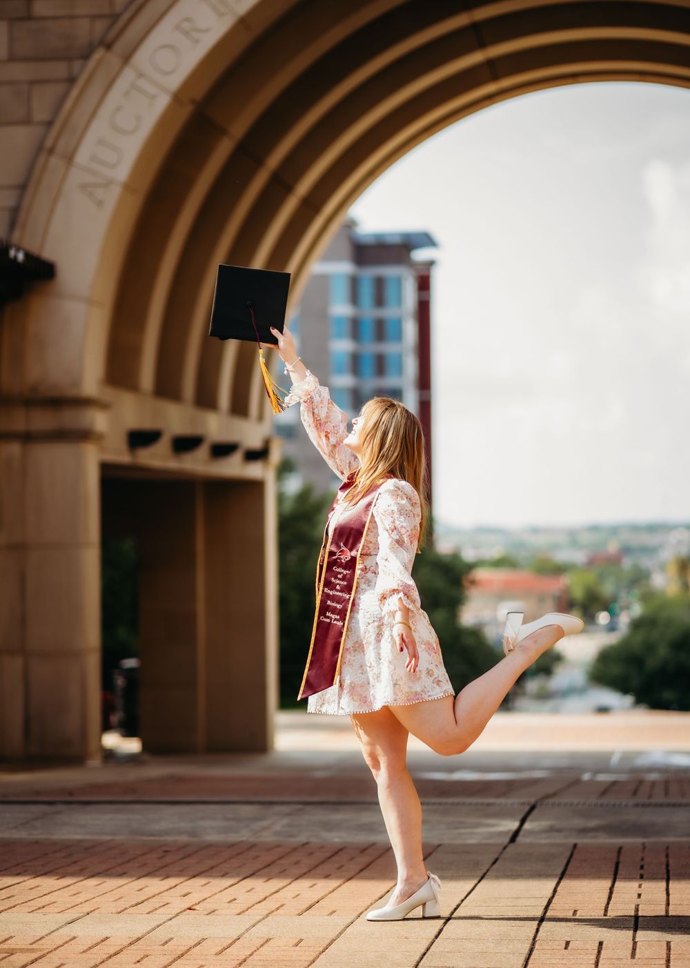 Graduate in a floral dress and graduation stole jumping joyfully under an archway.