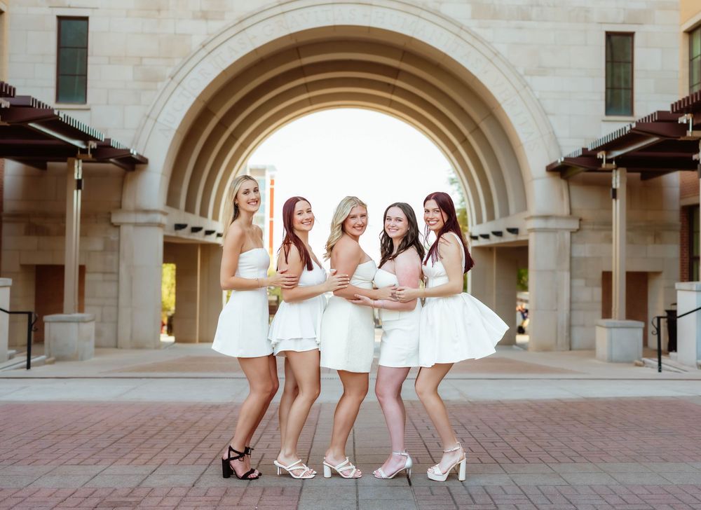 Group in matching white dresses pose together under an elegant stone archway.