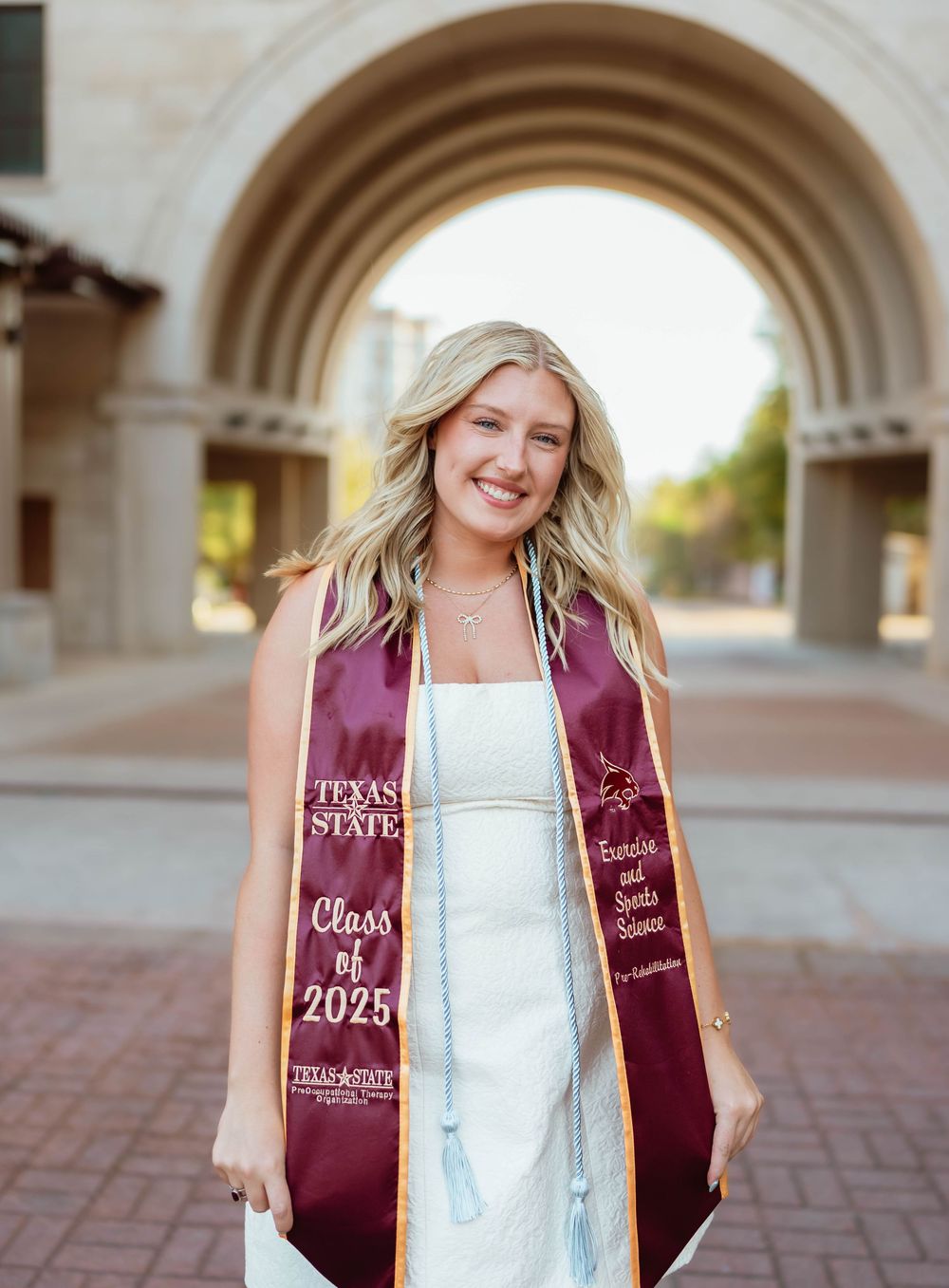 Graduate wearing Texas State graduation stole and dress beneath decorative campus archway.