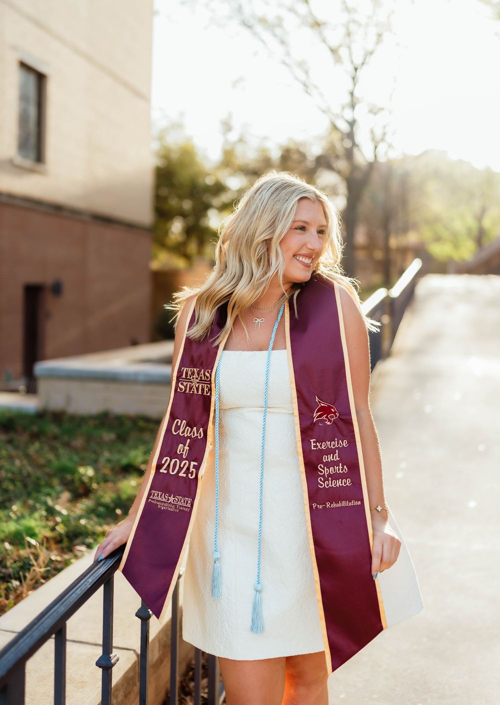 Graduate in a white dress and maroon graduation stole stands smiling on outdoor steps in sunlight.