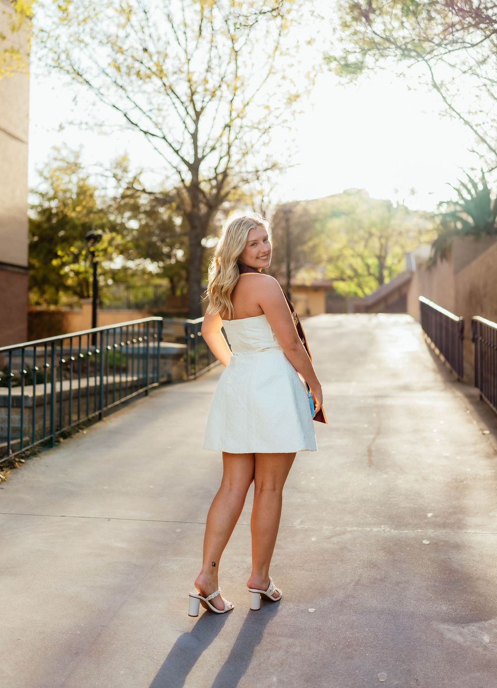 Graduate poses in white dress on sunny walkway at college campus during golden hour.