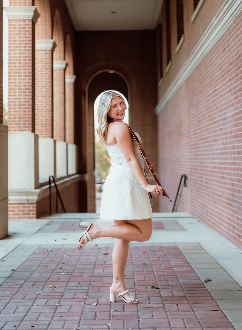 Person in a white summer dress poses playfully in an archway hallway with brick walls and arched architecture.