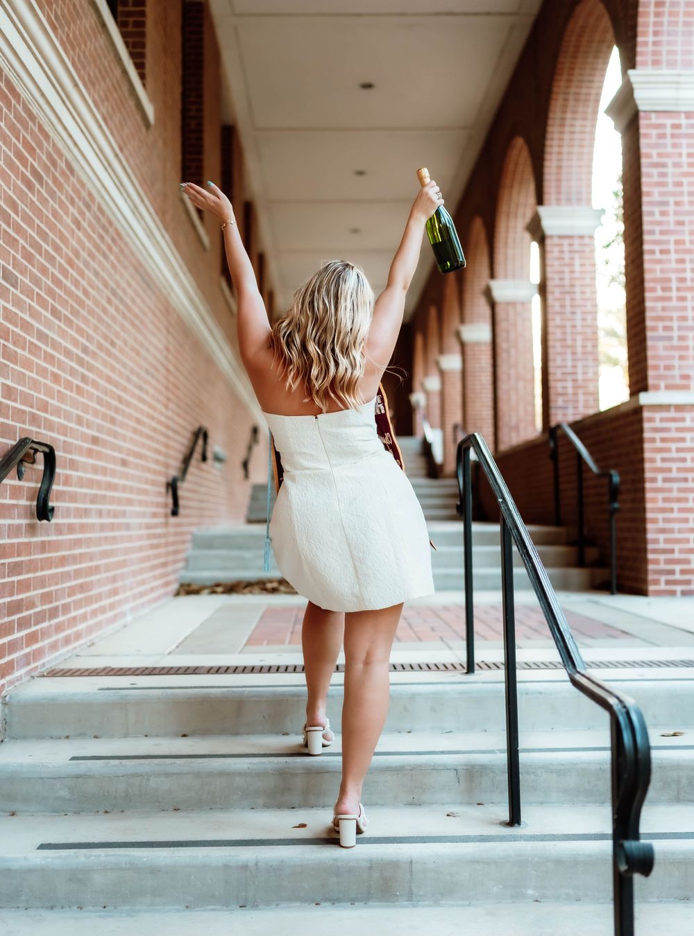 Graduate celebrates with arms raised in white dress on brick-lined campus walkway.