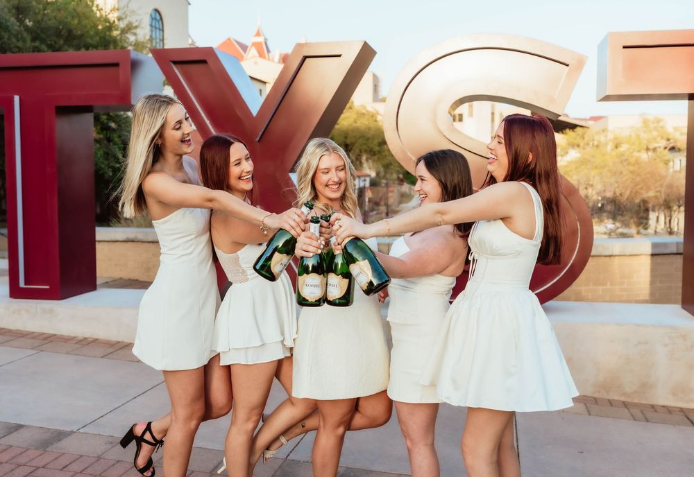 A joyful moment captured outdoors with five women in white dresses celebrating together near vintage signage.