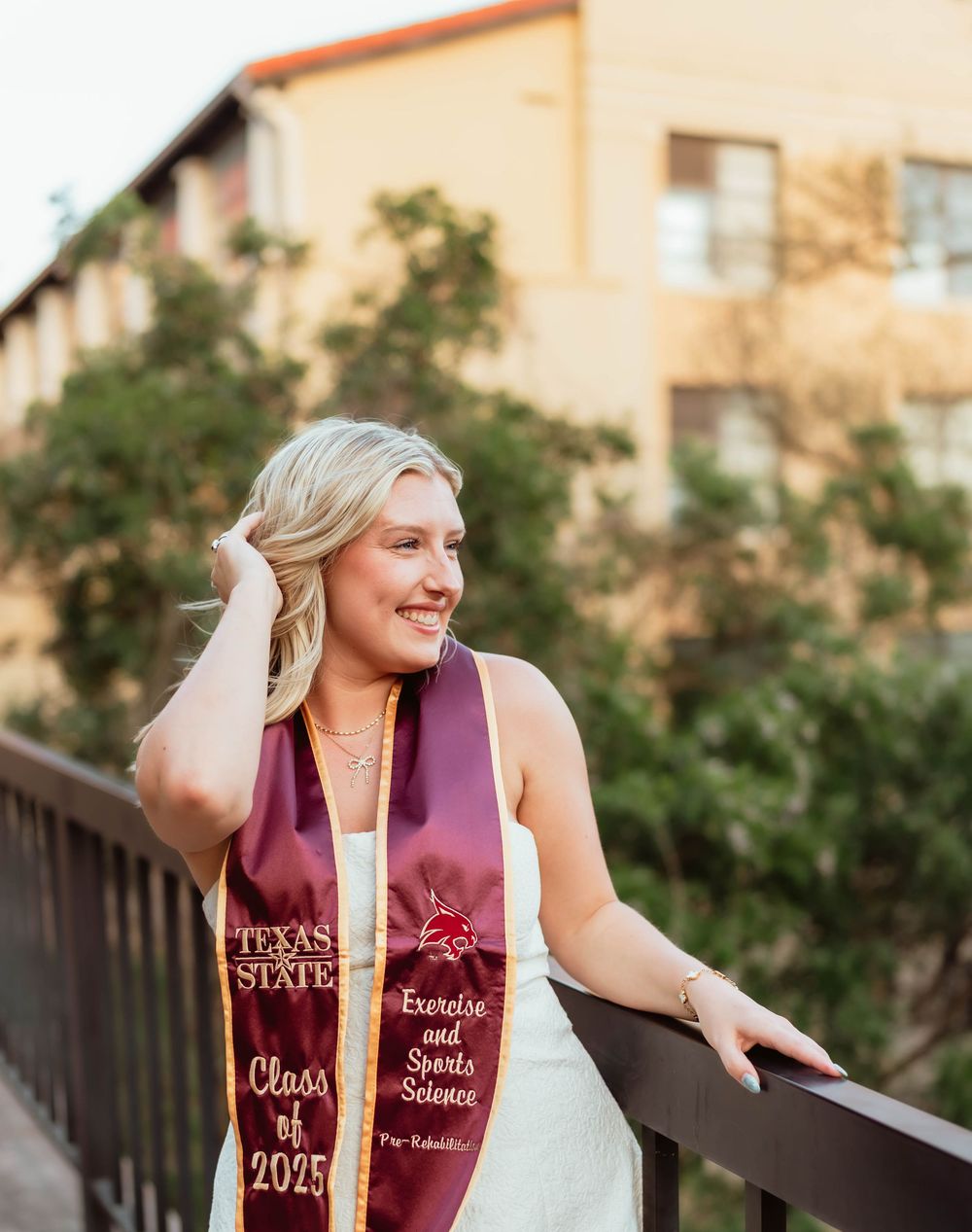 Graduate wearing burgundy Texas State graduation stoles stands outdoors in white dress for graduation photos.
