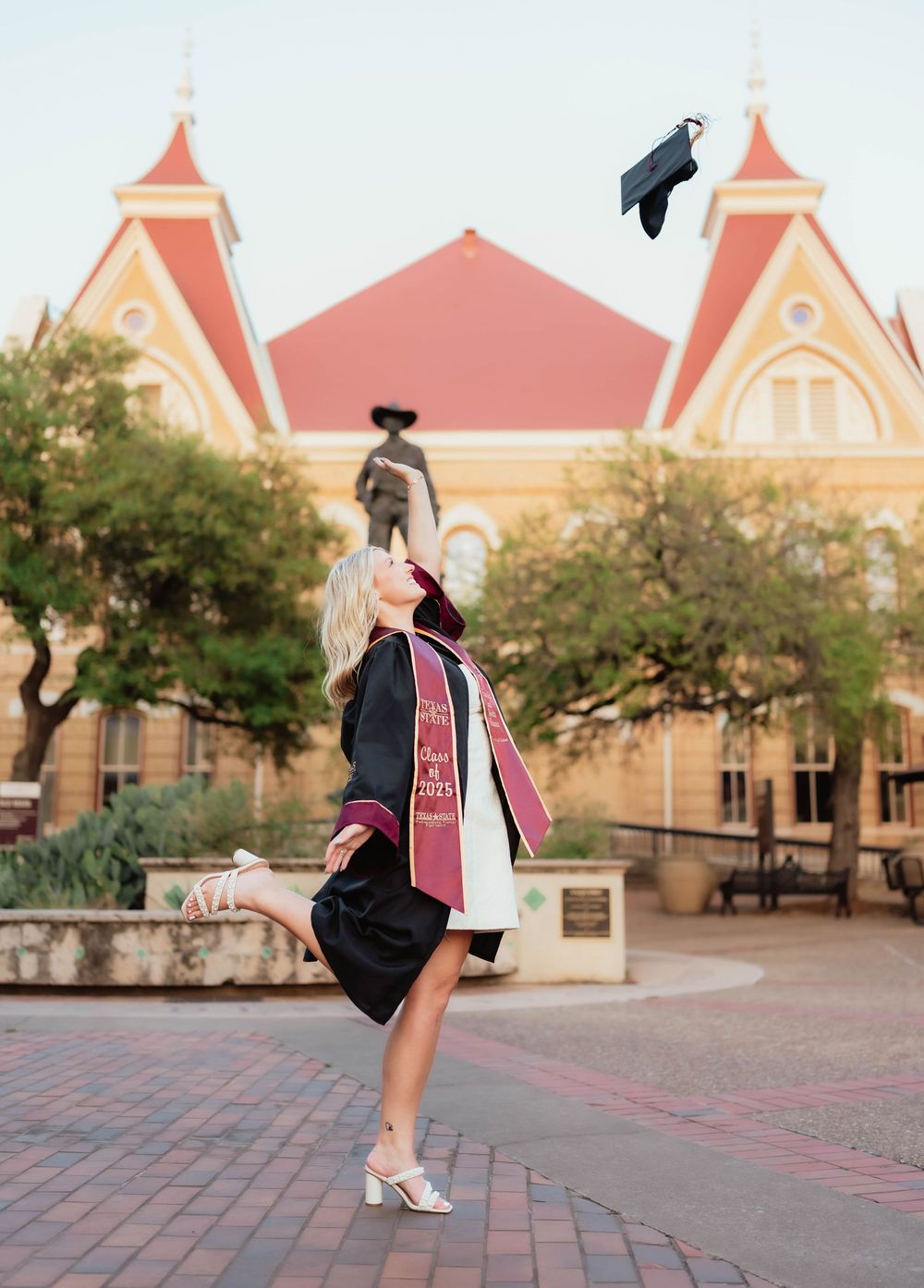 Graduate tosses cap in front of pink Victorian style campus building during graduation celebration.