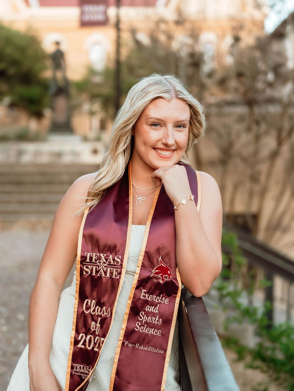 Graduate wearing maroon Texas State graduation stole and white sash stands outdoors during commencement ceremony.