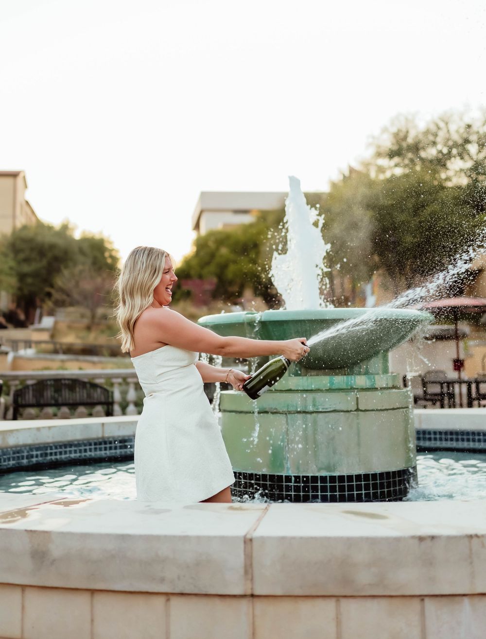 A graduate in a white dress celebrates by a fountain at Texas State University.