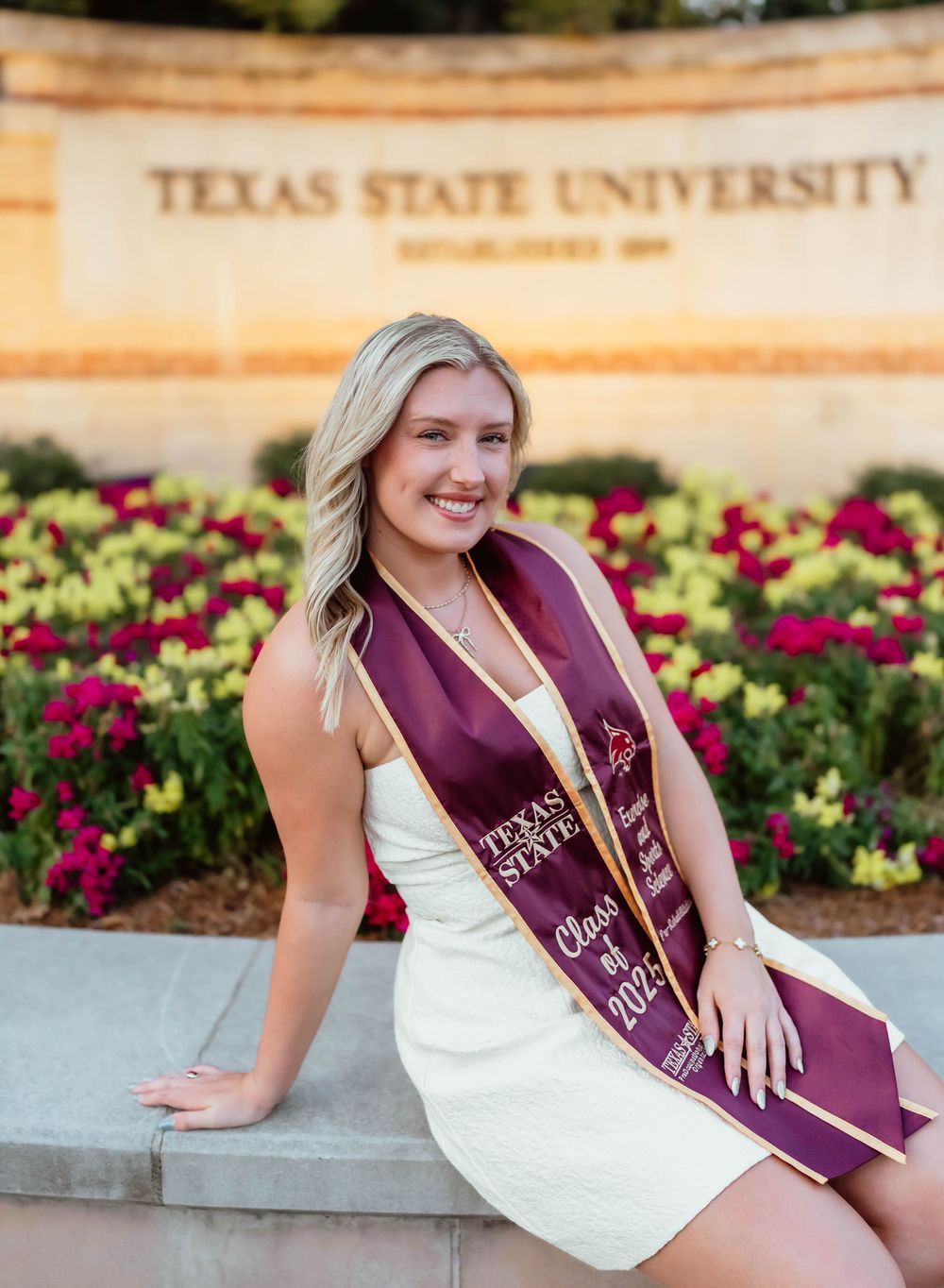 Graduation photos taken at the Texas State University entrance with maroon and gold flowers.
