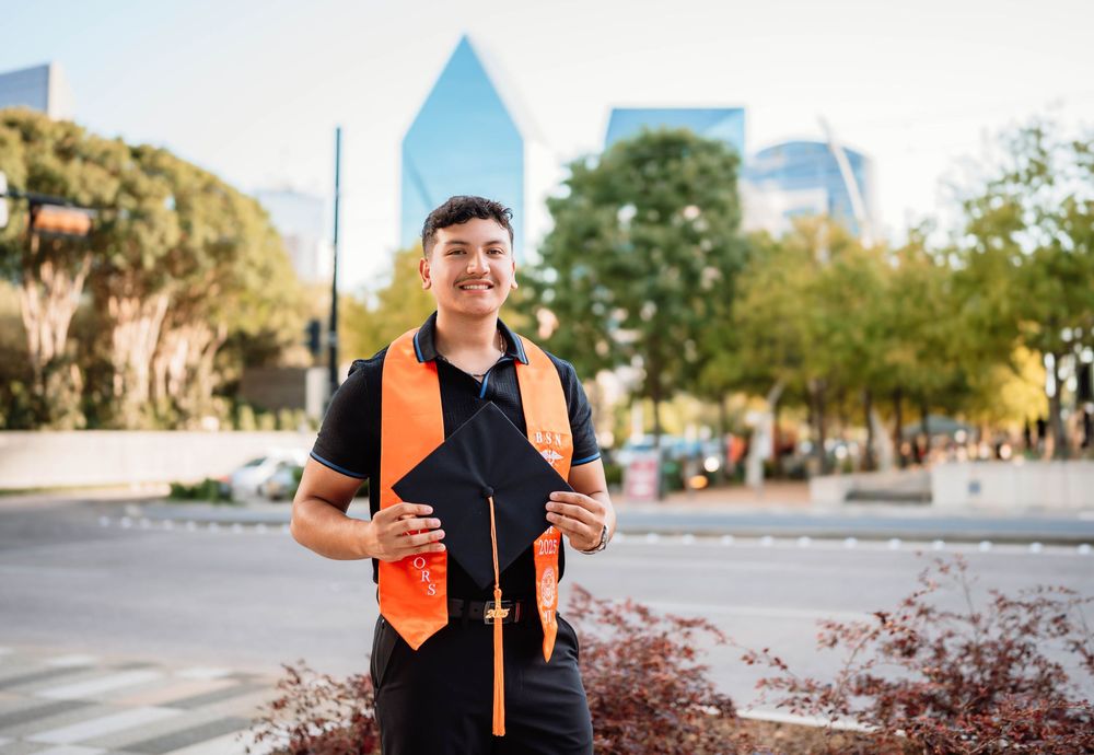 A person wearing a black t-shirt and orange vest stands holding a walking stick in an urban area with buildings behind.