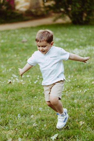 Child playing and jumping on grassy lawn wearing white shirt and shorts.