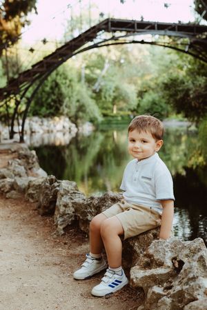 Child sitting on rocks by a serene pond in casual summer attire.