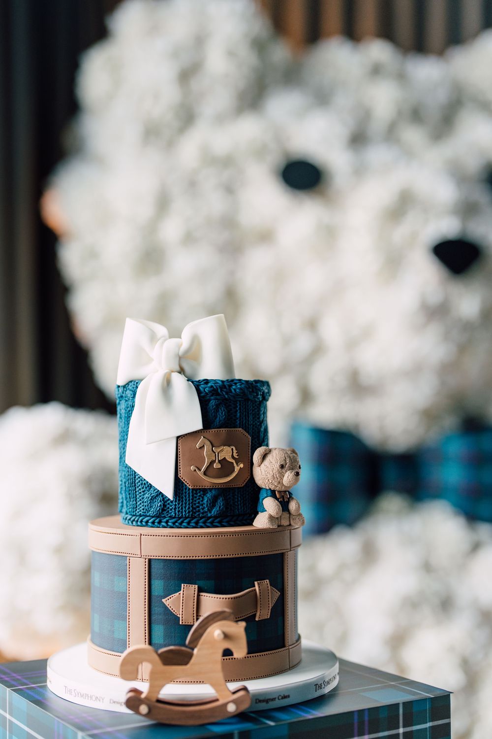 Blue and white baby shower cake decorated with rocking horse motif and bow on top, set against cloudy backdrop.