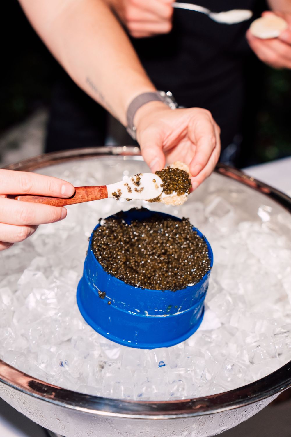 Hands carefully arranging black caviar on a blue plate set over crushed ice on a silver serving platter.