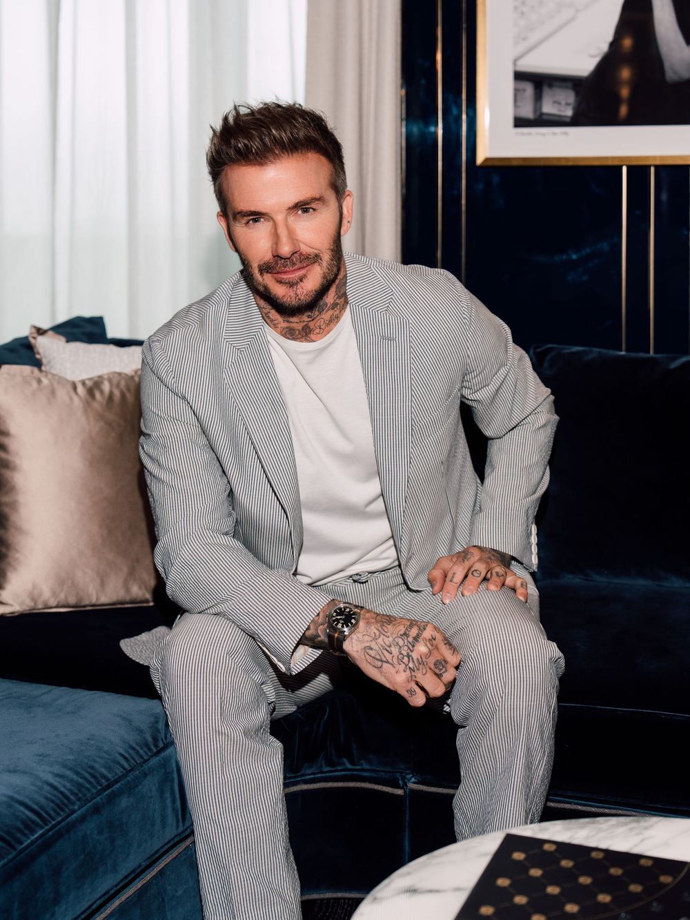 David Beckham wearing a light gray suit and white t-shirt sits on a blue velvet couch in an elegant setting at the Londoner Hotel in Macau