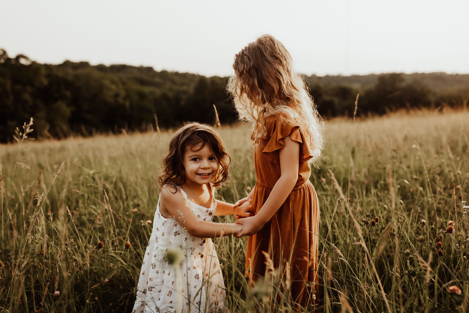 Two young sisters in summer dresses hold hands and smile together in a sunlit golden grass meadow.