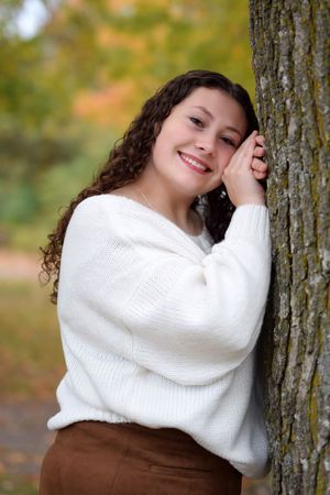 Person in cozy white sweater leans against tree trunk during autumn photoshoot.